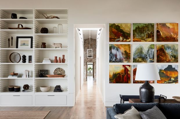 Bright, refined interior view from a living room toward a long hallway. Built-in white display shelving fills the left wall, while a dark sofa, side table, and table lamp occupy the foreground. A large grid of colourful abstract artworks forms a feature wall on the right. The hallway has light timber flooring, white walls, and a high ceiling with a visible pendant light and distant window.