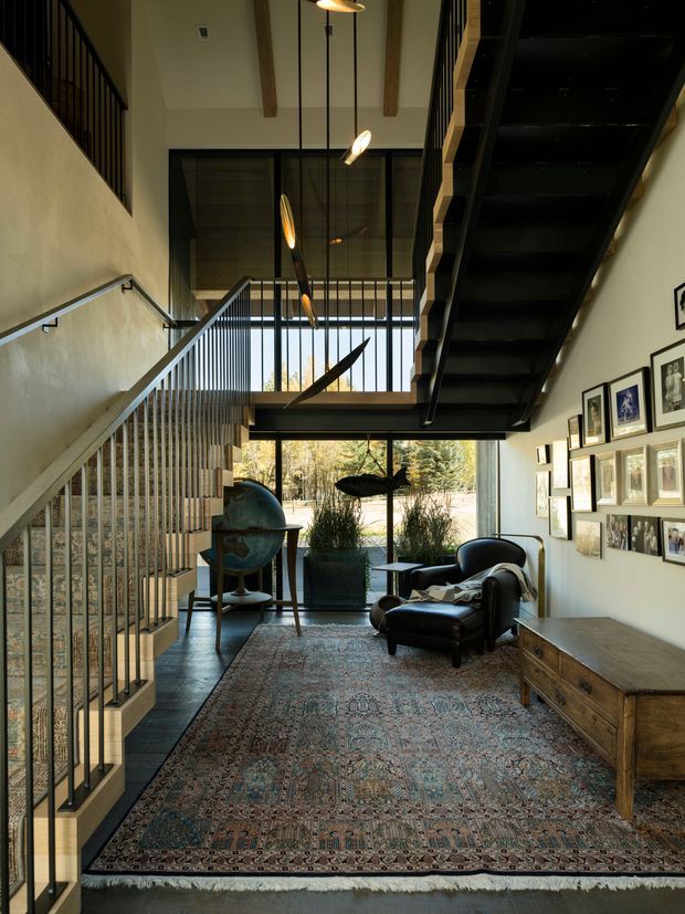 A tall, narrow double-height interior circulation and living space with a prominent black metal stair and mezzanine. The room features exposed timber ceiling beams, black-framed glazing, metal balustrades, a leather lounge chair, a vintage globe, framed artwork, a timber console, and a large patterned rug. Natural light enters through the full-height rear glazing, creating a warm, gallery-like atmosphere.
