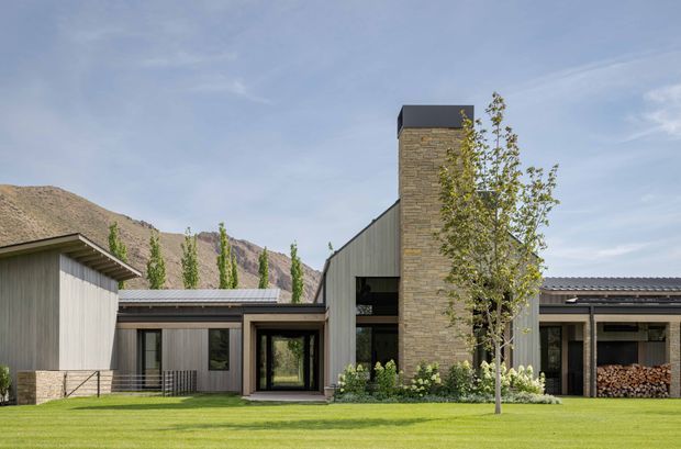 A contemporary single-storey residential building viewed from the landscaped front lawn. The composition features several low horizontal wings with grey metal pitched roofs, vertical grey timber-like cladding, extensive dark-framed glazing, and a prominent tall stone-clad chimney or tower with a dark cap. A neat lawn, young deciduous tree, planted beds, and stacked firewood frame the building, with rugged mountain terrain visible in the background.