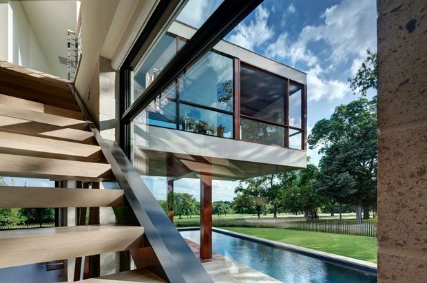 Architectural photograph viewed from inside a modern multi-level home, showing an open floating timber staircase with a dark metal stringer, extensive black-framed glazing, and a projecting glass-walled upper-level room. A narrow swimming pool, manicured lawn, mature trees, and blue sky are visible beyond the glazing. The design emphasises strong indoor–outdoor connection, exposed structural elements, and a high degree of transparency.