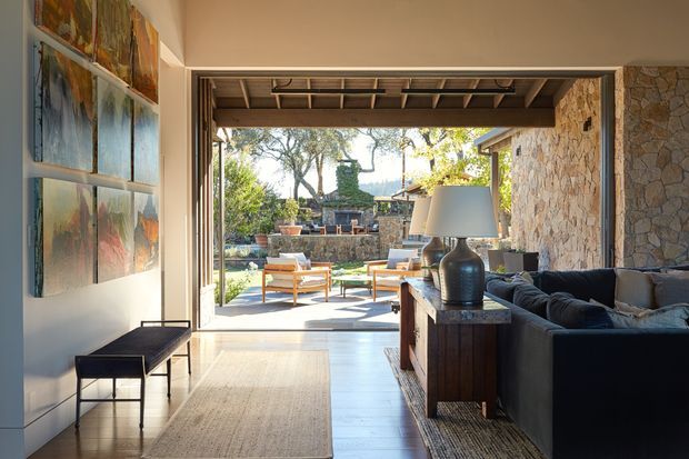 A refined mid-century-inspired living room opening through fully glazed sliding doors to a sunny outdoor terrace and landscaped garden. The interior features pale timber flooring, a neutral rug, dark upholstered seating, a timber console, sculptural lamp, and a large abstract artwork display. Exposed timber beams continue across the covered outdoor area, while a textured natural-stone wall forms a strong architectural feature. The space is bright, warm, and visually connected to the outdoors.