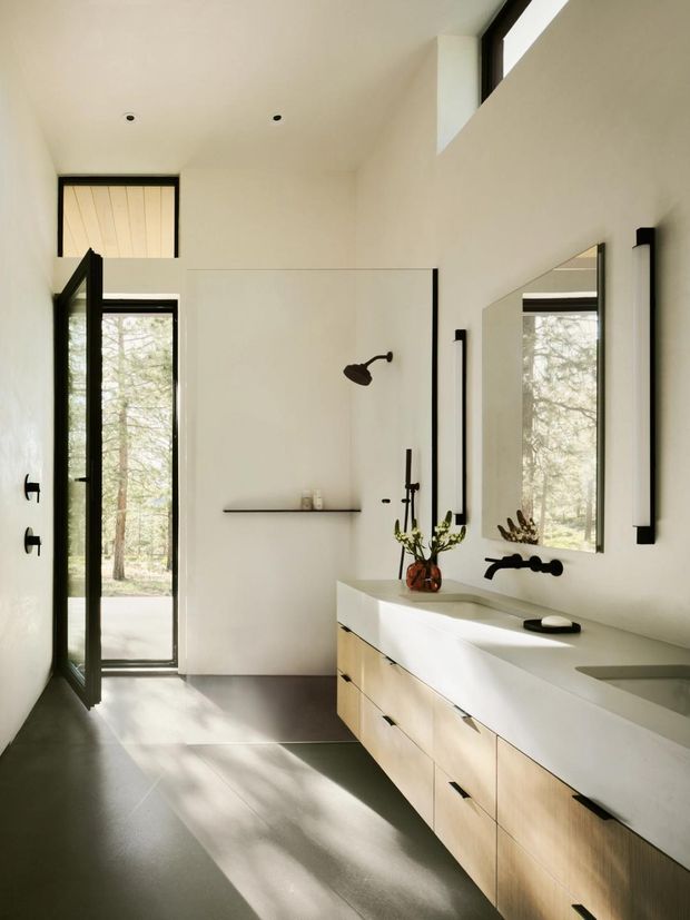 A bright, minimalist bathroom with a long double vanity, pale timber cabinetry, a white solid-surface benchtop, wall-mounted black fixtures, and a large frameless mirror. A glass shower enclosure and full-height glazed door open toward a wooded exterior. The room has high ceilings, clerestory glazing, recessed downlights, and a restrained black, white, and light-wood palette.