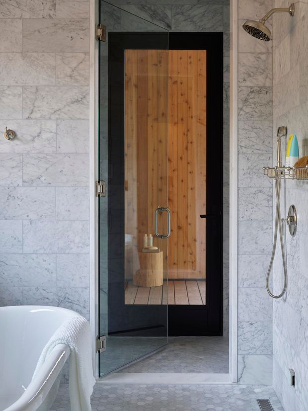 A refined bathroom with pale veined marble wall tiling, a glass-enclosed shower, and a partial freestanding white bathtub in the foreground. The shower includes brushed metal fixtures, a handheld hose, and a wall-mounted shelf. Through the glass, a warm timber door and timber-floored adjoining area are visible, creating contrast with the cool marble and dark framing. Small-format pale hexagonal tiles cover the bathroom floor.