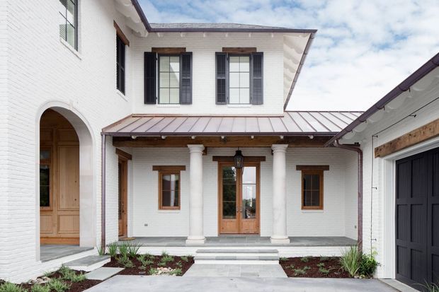 Exterior courtyard-facing view of a refined two-storey residence with white-painted brick walls, timber-framed windows and doors, dark shutters, classical columns, arched entry, and a standing-seam metal roof. Paved paths, planting beds, and an attached garage frame the central entrance.