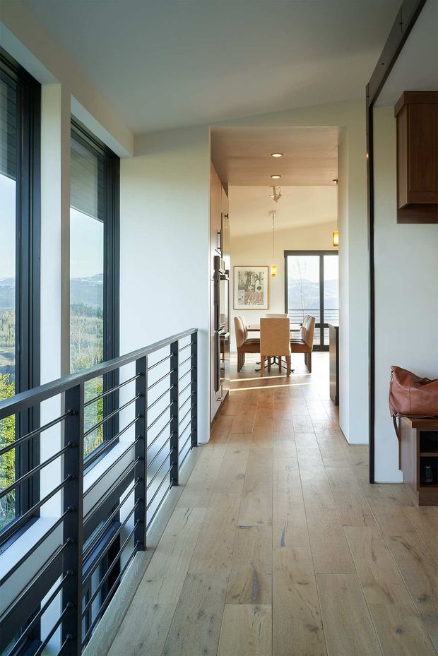 A bright interior hallway or landing with wide pale timber floorboards, white walls, and large dark-framed windows overlooking a wooded mountainous landscape. A black metal guardrail lines the window edge, and the corridor opens toward a dining area with a timber table and upholstered chairs. Built-in dark timber cabinetry and a leather chair are visible at the right edge. The space has a clean contemporary feel with recessed and pendant lighting.
