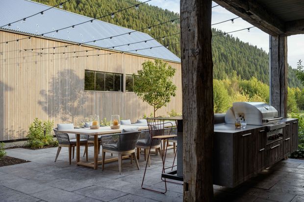 A covered outdoor entertaining area opens onto a paved courtyard beside a timber-clad building. The space includes a dining table with chairs, an outdoor kitchen with built-in stainless-steel barbecue and dark timber cabinetry, overhead festoon lighting, and a surrounding forested mountain setting. The architecture uses vertical timber cladding, a simple pitched metal roof, and substantial rustic timber posts and beams.