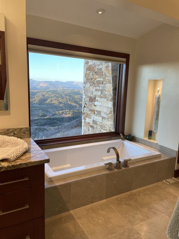 A luxurious bathroom with a large built-in bathtub positioned beneath a picture window overlooking a mountainous landscape. The room features warm beige stone or porcelain tile flooring and tub surround, dark wood vanity cabinetry, a polished stone countertop, dark wood window trim, recessed lighting, and a sculptural wall niche with decorative objects. A stone-clad exterior wall is visible through the window.