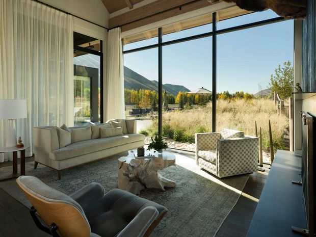 A refined living room with expansive floor-to-ceiling glazing overlooking a dry meadow and mountain landscape. The space features a pale upholstered sofa, patterned armchairs, a large rug, a sculptural natural wood coffee table, dark built-in cabinetry, sheer curtains, and an exposed timber ceiling structure. Strong indoor-outdoor connection and abundant natural light define the room.