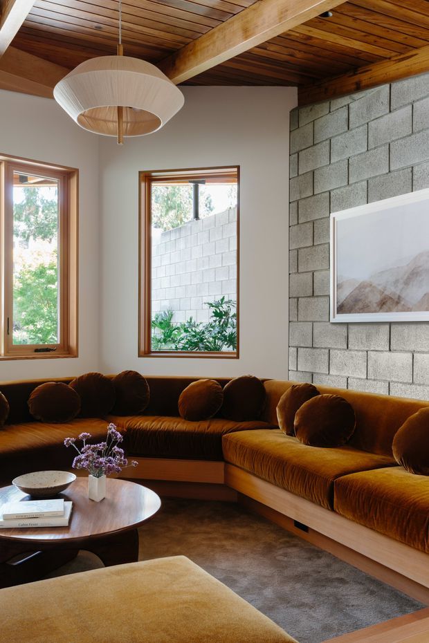 Warm, richly furnished living room with a large caramel-brown velvet sectional sofa, round wooden coffee table, upholstered ottoman, and soft area rug. The room features exposed timber ceiling boards and beams, natural timber window frames, a grey concrete block feature wall, and a sculptural pendant light. Large windows bring in daylight and greenery, creating a cozy mid-century-inspired atmosphere.