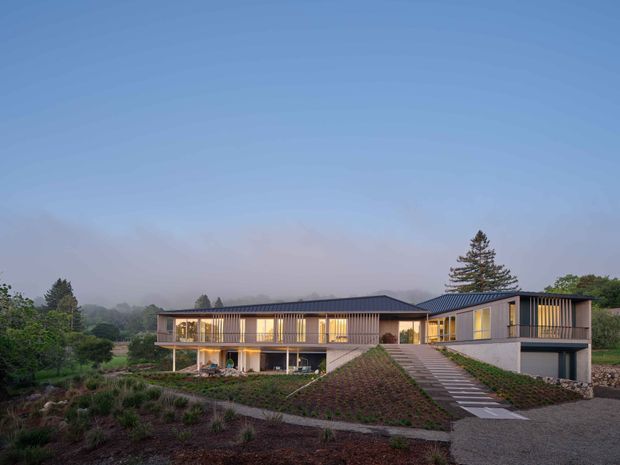 A wide dusk exterior view of a large contemporary single-storey home set within landscaped grounds. The residence has a low, elongated form with dark metal gable roofs, extensive floor-to-ceiling glazing, warm illuminated interiors, vertical timber-like screening, and a prominent stepped concrete path leading up to the entrance. The surrounding garden includes planted terraces and a gravel driveway, with mature trees in the background.