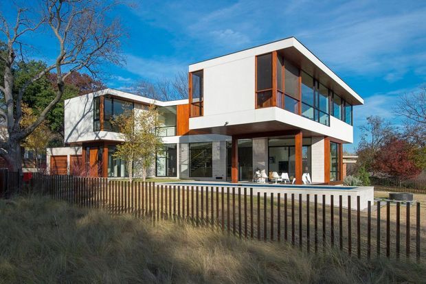 Exterior view of a contemporary multi-level residence with crisp white rendered walls, strong horizontal rooflines and cantilevered upper volumes. Extensive floor-to-ceiling glazing and warm timber-framed windows and posts create contrast against the white facade. The home sits behind a vertical dark metal or timber slat fence, with landscaped planting and mature deciduous trees surrounding it.