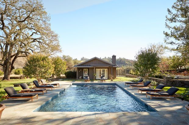 A landscaped outdoor pool area featuring a long rectangular swimming pool, paved stone surrounds, and multiple modern timber-and-cushion sun loungers. A single-storey, symmetrical gabled pavilion with vertical timber cladding and a central chimney sits beyond the pool, framed by mature trees, hedges, and a low stone retaining wall. The scene is sunny, spacious, and resort-like.