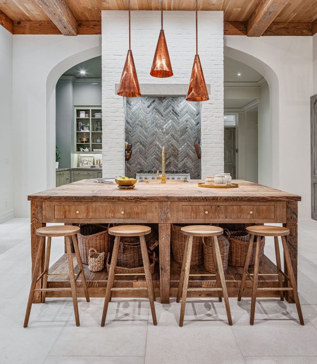 A warm, richly detailed kitchen centered on a large rustic timber island with drawers, woven basket storage, and five timber stools. Three copper pendant lights hang above the island, while a white-painted brick chimney breast frames a herringbone-patterned metallic backsplash and cooking area. Arched openings reveal built-in cabinetry and shelving on either side. Exposed timber ceiling beams and pale large-format floor tiles complete the refined rustic interior.