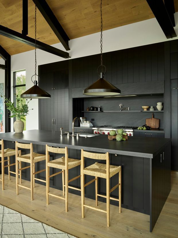 A sophisticated open-plan kitchen dominated by matte black vertical-panel cabinetry and a large dark island with seating for four. The kitchen features a dark stone-look benchtop, integrated shelving, black appliances and tapware, warm timber flooring and ceiling, exposed dark timber beams, and two oversized bronze pendant lights. Natural light enters through tall windows, while woven timber bar stools add warmth and texture.