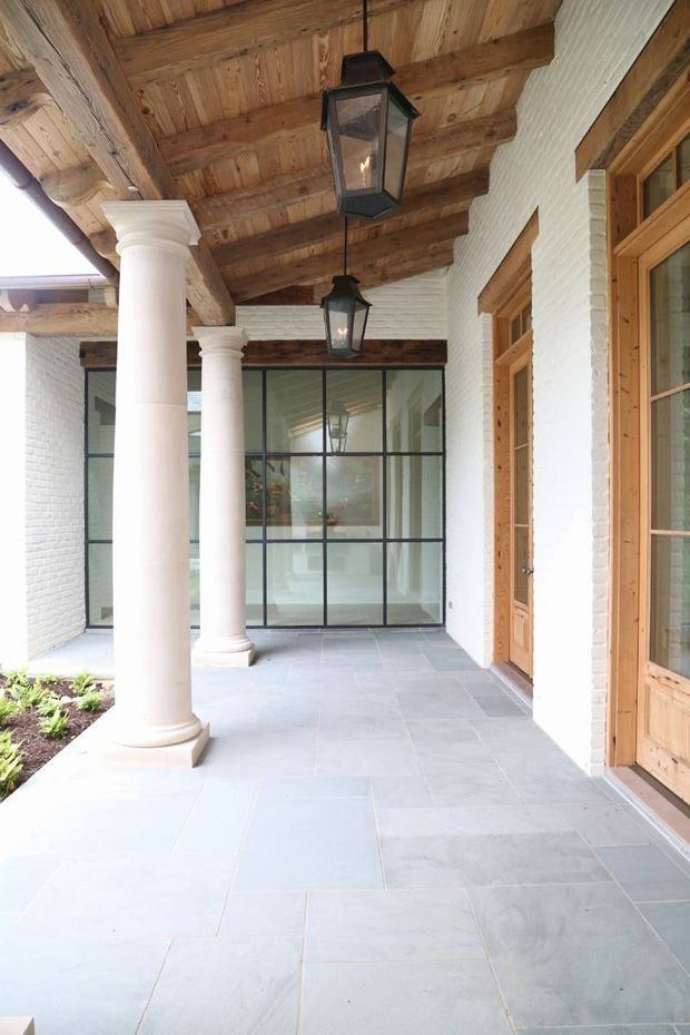 Covered exterior colonnade or veranda with white painted brick walls, classical round columns, a heavy exposed timber-beam ceiling, hanging lanterns, large grey stone floor tiles, black-framed glazed doors, and warm timber-framed entry doors. The space has a refined traditional character with a bright, airy courtyard-like feel.