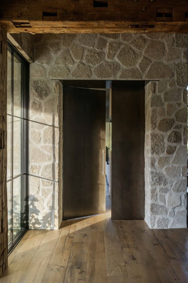 A rustic interior entryway featuring a heavy exposed timber ceiling beam, rough irregular natural-stone walls, tall black-framed glazing on the left, wide timber floorboards, and a pair of tall dark timber pivot doors opening into another space. The image is warmly lit with a strong emphasis on natural materials and handcrafted detailing.