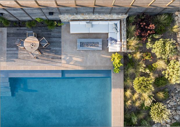 Top-down aerial view of a landscaped outdoor entertaining area with a swimming pool, adjoining paved terrace, timber deck, outdoor dining setting, and built-in lounge with a rectangular fire feature. Dense ornamental planting and rock landscaping border the space, while a timber privacy screen lines the upper edge.