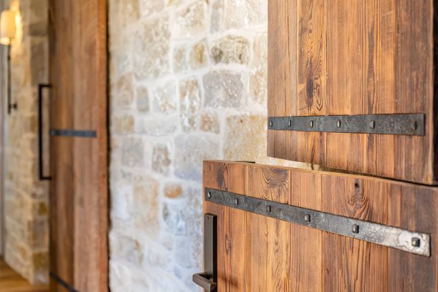 Close-up interior view of rustic timber doors with prominent dark metal strap hinges and handles, set against a pale irregular stone masonry wall. The warm wood grain, heavy hardware, and textured stone create a traditional farmhouse or barn-inspired character.