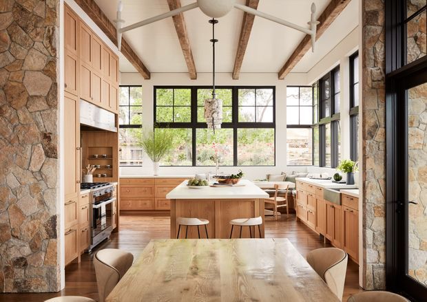 A bright, spacious open-plan kitchen and dining area with extensive natural light, warm timber cabinetry, a central island, and a large dining table. The room features exposed timber ceiling beams, a stone-clad structural wall, dark-framed floor-to-ceiling glazing, stainless-steel appliances, and a neutral warm palette. The kitchen opens visually toward a landscaped outdoor setting.
