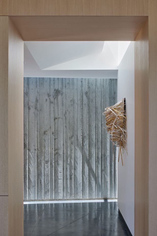 A minimalist interior passage or entry space framed by light timber wall and ceiling joinery. The room features a dark polished floor, a vertically ribbed metallic grey feature wall, white sculptural ceiling geometry, and a small abstract natural-fibre wall artwork. The composition is calm, refined, and strongly architectural, with daylight entering from above.