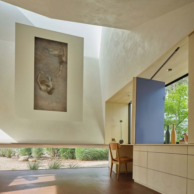 A refined, light-filled interior with a sculptural curved plaster ceiling and walls, a large framed artwork, and a compact built-in study or workspace. Pale timber cabinetry with flush drawers and a tall blue panel frames the desk area, which includes a simple timber chair and minimal desk accessories. Full-height glazing opens to a landscaped garden, while warm timber flooring and filtered daylight create a calm, contemporary atmosphere.