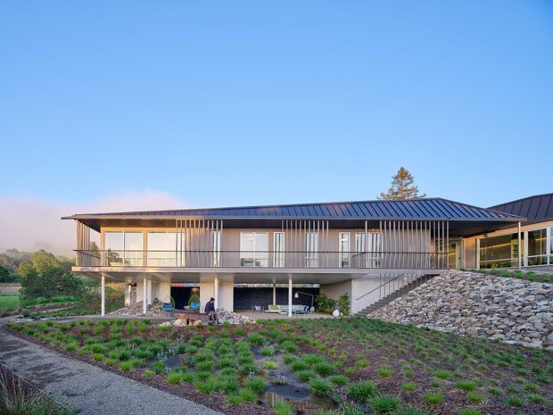 A contemporary multi-level residential building set into a landscaped sloping site. The long low-slung structure has a dark standing-seam metal roof, extensive glazing, balconies, slender vertical screening, exposed structural supports, and a stone retaining wall. Exterior stairs connect the upper living level with the lower ground-level area, while planted beds and gravel landscaping surround the house.
