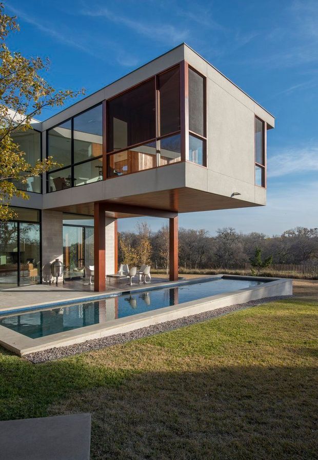 Exterior view of a contemporary multi-storey residence with a projecting upper volume, extensive floor-to-ceiling glazing, concrete-toned walls, dark timber soffit, and reddish timber or metal framing. A long rectangular swimming pool and paved terrace sit beside the house, with landscaped lawn and trees beyond.