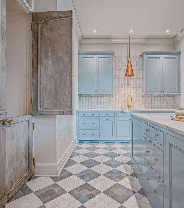 A refined kitchen with pale blue raised-panel cabinetry, a large central island, white countertops, and a tiled backsplash. The floor features a geometric checkerboard pattern in soft blue, grey, beige, and white. A warm copper pendant hangs above the island, while a distressed timber pantry door and matching lower cabinetry add character. The space has decorative ceiling mouldings, recessed lighting, and a bright, elegant traditional fit-out.