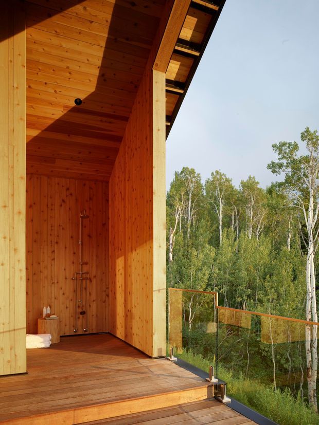 A timber-lined outdoor shower enclosure opens onto a raised timber deck with frameless glass balustrading and views into a wooded landscape. The shower area has warm vertical wood walls, a timber ceiling and exposed structural post, creating a sheltered indoor-outdoor bathing space.
