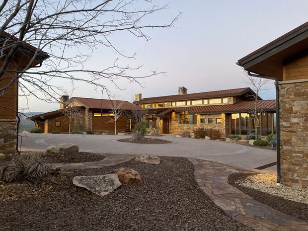 A broad exterior view of a low-rise lodge-style building complex arranged around a landscaped courtyard. The buildings feature natural stone walls, dark timber cladding, warm wood soffits, and low-pitched reddish metal roofs. Stone-paved walkways, gravel garden beds, large boulders, and leafless trees frame the scene, with warm interior lighting visible through the windows.