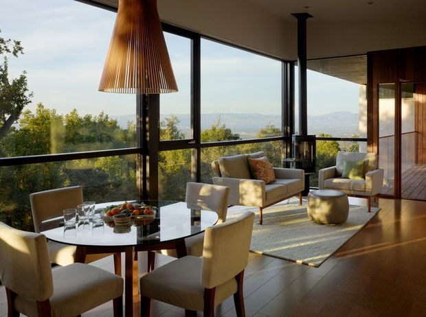 A sophisticated open-plan living and dining area with expansive floor-to-ceiling glazing overlooking a wooded landscape and distant mountains. The room features a round glass dining table with upholstered chairs, a compact seating area with armchairs and a sofa, a large woven rug, warm timber wall panelling and doors, dark slim window frames, and a sculptural timber pendant light. Natural sunlight creates a warm, refined atmosphere.