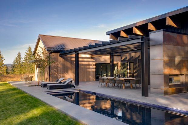 Exterior architectural view of a contemporary home at dusk, featuring a dark metal flat-roofed pavilion with exposed timber rafters, a covered outdoor dining terrace, a reflecting swimming pool, and a contrasting pitched-roof wing clad in warm timber or metal. The setting includes landscaped lawn and trees, with strong indoor-outdoor connection and warm architectural lighting.