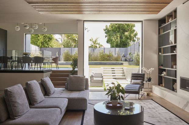 A refined open-plan living room with a large grey sectional sofa, round black coffee table, built-in shelving and media cabinetry, and full-height glazing opening to a landscaped courtyard. A dining area is visible to the left, while the ceiling features a linear timber-slat detail and the interior uses a restrained neutral palette.