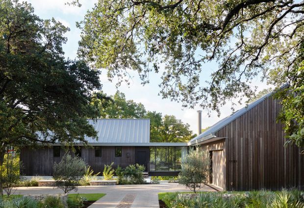A low-rise contemporary residential complex arranged around a landscaped courtyard. The buildings feature dark vertical timber cladding, simple pitched and folded metal roofs, extensive glazed connecting sections, and a paved central pathway framed by established trees and drought-tolerant planting.