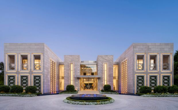 A symmetrical, grand contemporary house facade viewed from the front. The building has two matching flat-roofed wings clad in pale stone or concrete panels, tall vertically proportioned windows with dark frames, decorative patterned screens, and a recessed central entrance with extensive glazing. A circular reflecting pool and formal clipped planting sit in the forecourt under a clear blue sky.