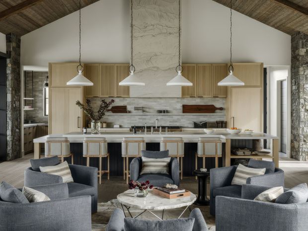 Open-plan kitchen and living room with a large central island, five bar stools, layered pendant lighting, and a surrounding seating area. The space features exposed rustic timber ceilings, rough stone columns and walls, light horizontal wall tiling, fluted timber cabinetry, a tall marble-look chimney feature, and dark upholstered lounge chairs arranged around a round coffee table.