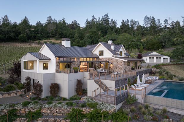 A multi-level contemporary residence set into a landscaped, wooded and vineyard-like hillside. The home has white rendered walls, dark standing-seam pitched roofs, stone-clad exterior walls, extensive glazing, multiple terraces and decks, and a swimming pool beside the lower outdoor entertaining area. A detached garage or outbuilding is visible to the right.