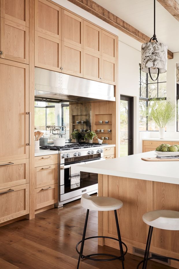 Warm, light-filled kitchen with extensive oak cabinetry, a large white island with stools, stainless-steel range and hood, open display shelving, timber flooring, and large black-framed windows. Exposed rustic timber ceiling beams and a sculptural pendant add character to the otherwise refined, contemporary space.