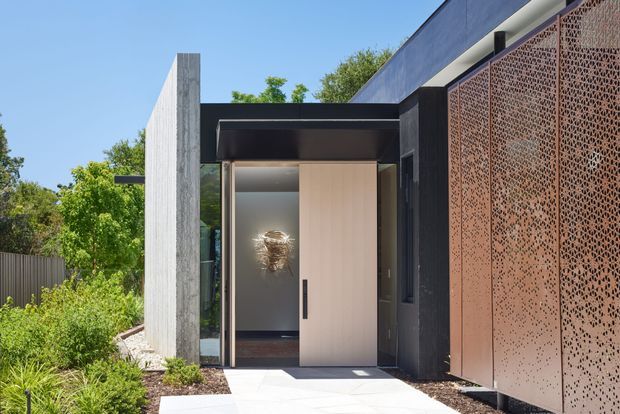Modern residential entry facade with a sheltered recessed doorway, pale timber or wood-look pivot door, dark metal framing, textured concrete wall, and a perforated rust-toned metal screen. A landscaped garden and paved path lead to the entrance.