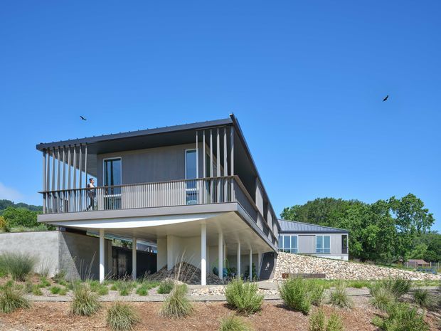 Exterior photograph of a contemporary elevated two-storey residence with a long projecting upper level, extensive vertical slatted screening, broad balcony, slender columns, and large glazed openings. The building has muted grey and white finishes, a low-pitched metal roof, and stone-clad lower walls, set within a landscaped dry garden.