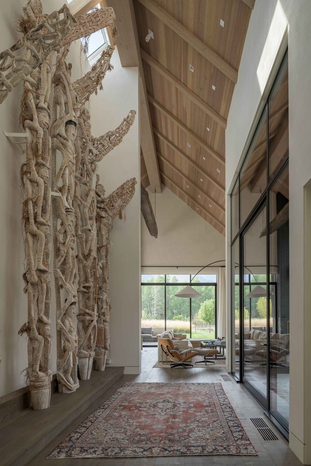 A tall, refined residential interior hallway or entry gallery with a dramatic double-height volume. The space features a warm timber-lined pitched ceiling with exposed structural beams, pale plaster walls, full-height black-framed glazing and doors, and a large sculptural carved wood installation along the left wall. A patterned area rug leads toward a bright living area furnished with a leather lounge chair and ottoman, sofa, and pendant light. The glazing reflects the interior and opens the room visually toward landscaped grounds.