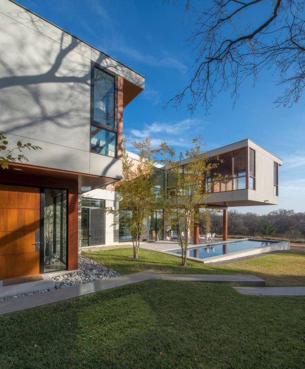 Exterior view of a contemporary multi-wing residence arranged around a landscaped courtyard and rectangular swimming pool. The home features crisp box-like volumes, pale smooth facade panels, extensive floor-to-ceiling glazing, warm timber entry doors and vertical timber-toned screening, with deep projecting upper floors and slender supports. Concrete paths, lawn, ornamental trees, and a blue-sky setting frame the architecture.