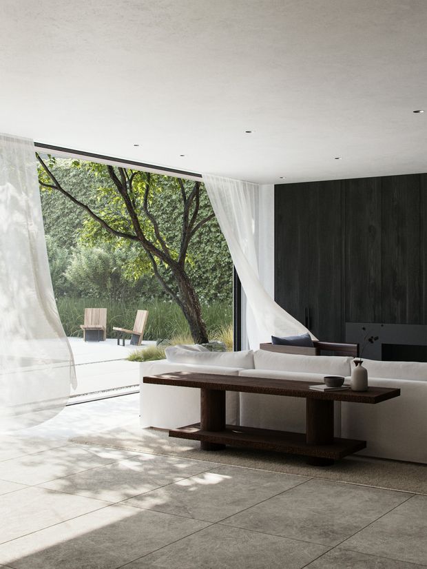 Bright contemporary living room with a low white ceiling, expansive floor-to-ceiling glazing, sheer white curtains, a white sectional sofa, and a substantial dark timber console table. A dark vertically panelled feature wall with a minimalist fireplace anchors the room, while mature greenery is visible through the open glazing. Large-format pale stone or concrete-look floor tiles create a calm, natural indoor-outdoor connection.