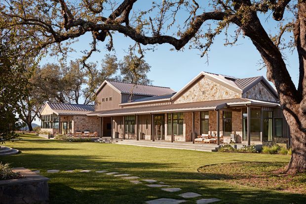 A sprawling single-storey residence set within a mature landscaped garden, featuring multiple connected pitched-gable volumes, extensive floor-to-ceiling glazing, stone-clad walls, and dark standing-seam metal roofs. A broad lawn and stepping-stone path lead toward covered outdoor terraces and glazed living spaces. Large established trees frame the contemporary country-style architecture.