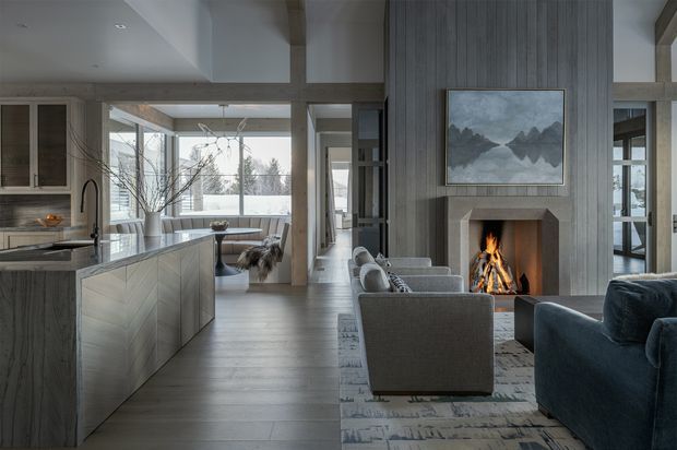 Open-plan contemporary interior combining a kitchen, dining area and living room. The space features a large island with grey chevron-patterned cabinetry, pale timber flooring, exposed timber structural beams, expansive glazing, and a central fireplace clad in vertically textured grey wall panelling. Neutral upholstered seating and a dark blue sofa surround the fireplace, creating a warm, refined atmosphere.