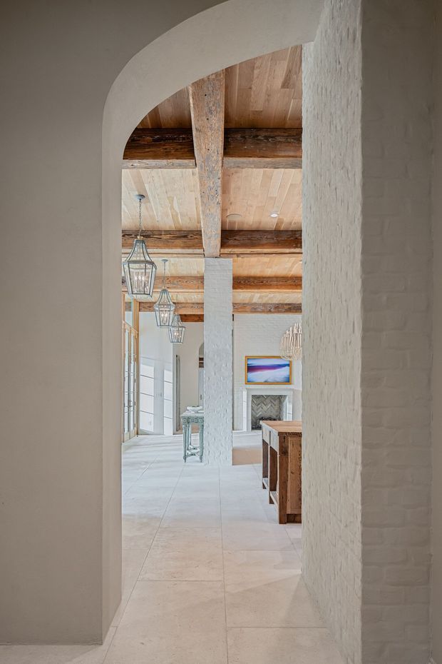 View through a rounded archway into a bright, open interior with white-painted masonry walls, exposed rustic timber beams, a pale timber ceiling, and large light-coloured stone or tile flooring. Lantern pendant lights line the space, with timber cabinetry or furniture visible to the right and a fireplace or decorative wall feature in the background. The design blends rustic structural elements with clean contemporary finishes.