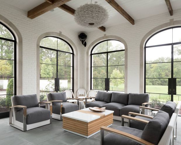 Bright, spacious living room with white painted brick walls, dramatic arched black-framed windows, exposed timber ceiling beams, and a contemporary seating arrangement around a layered wood-and-white coffee table. A sculptural woven pendant light creates a focal point, while the garden visible through the glazing provides a leafy outlook.