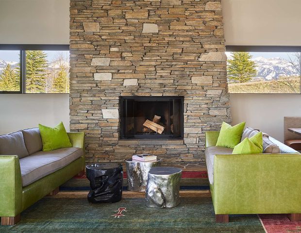 A contemporary living room centered on a substantial stacked natural-stone fireplace wall with a built-in firebox. Two lime-green and grey sofas face each other across a patterned area rug, with sculptural metallic and dark coffee tables between them. Narrow horizontal windows on either side of the fireplace frame a mountain landscape, while white walls and strong daylight create a bright, inviting interior.