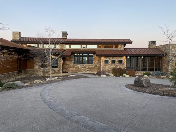 Single-storey residential building viewed from a broad curved driveway and landscaped courtyard. The facade combines rough natural stone masonry, dark timber wall sections, extensive dark-framed glazing, and a low-pitched standing-seam metal roof with several stone chimney elements. The restrained courtyard landscaping uses gravel, rocks, sparse shrubs, and leafless deciduous trees. The architecture has a rugged contemporary lodge or mountain-resort character.