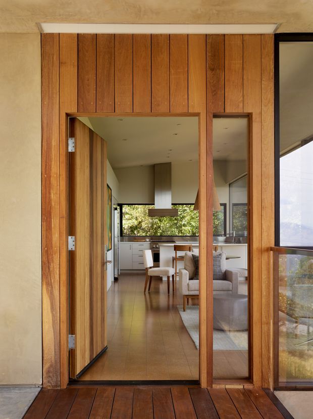 View from a timber deck through a tall timber-framed entry door into an open-plan interior. Vertical timber wall cladding surrounds the doorway, with a glazed sidelight and large windows revealing a dining area, kitchen cabinetry, and living room beyond. The interior uses warm timber floors and joinery, neutral walls, and abundant natural light.